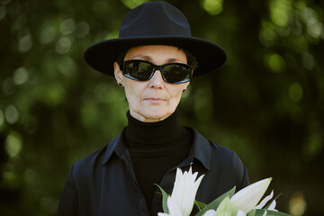 Mourning widow holding bouquet during burial service, she hiding her tears under sunglasses