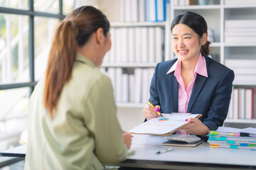 Two Asian businesswomen use laptop and tablet to collaborate in a productive conversation with colleagues while analyzing documents, financial graphs, working in the office.