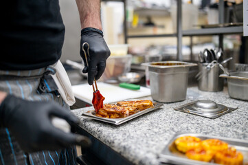 A chef in a professional kitchen is glazing cooked meat with sauce, focusing on the action of...