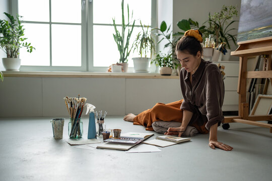 Creative woman designer painting with colors, comparing sketches in sketchbooks, sitting on floor in art workshop. Interested girl illustrator working with watercolor looks on picture thinks new ideas