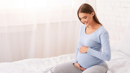 Enjoying Pregnancy. Pregnant Lady Touching Belly Smiling Sitting On Bed At Home In Morning. Free Space