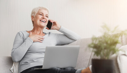 Cheerful elderly business lady talking by phone, sitting on sofa and using laptop, free space
