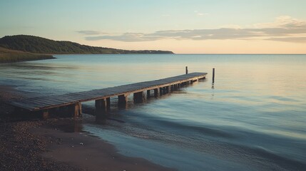 Obraz premium A peaceful beach with a small wooden pier extending into the water. The calm, reflective surface of the ocean and the soft light of sunset create a serene and contemplative scene