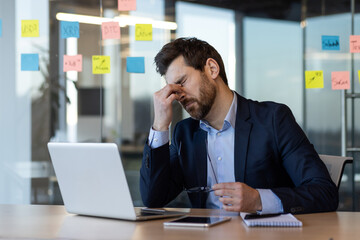 Businessman feeling stressed and overwhelmed, sitting at desk holding head in frustration. Office...