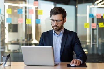 Businessman concentrating on laptop work in modern office setting, surrounded by colorful sticky notes on glass wall. Portrays focus, productivity, and analysis in business environment.