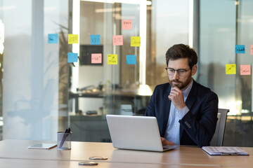 Businessman wearing suit concentrating on laptop work in contemporary office. He is surrounded by colorful sticky notes for strategic tasks. Ideal for business, planning, and technology concepts.