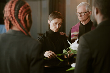 Senior pastor supporting mourning widow who laying white lily on black coffin