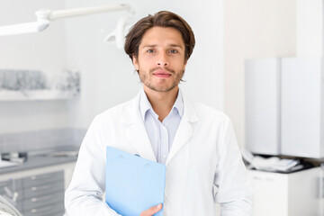 Fototapeta premium Visit my clinic. Handsome confident dentist holding folder over cabinet background, empty space