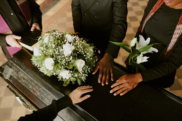 High angle shot of unrecognizable hands on black wooden coffin with white roses on top, pastor...