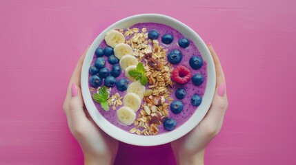 A beautifully arranged bowl of purple smoothie topped with fresh blueberries, banana slices, granola, and mint, held against a vibrant pink background.