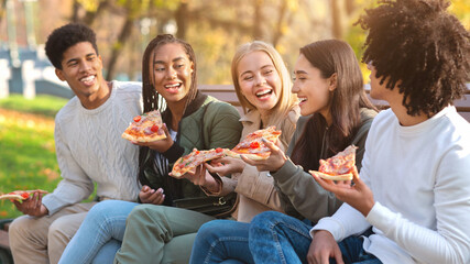 Fototapeta premium Hungry laughing international teenagers enjoying pizza while spending time in park