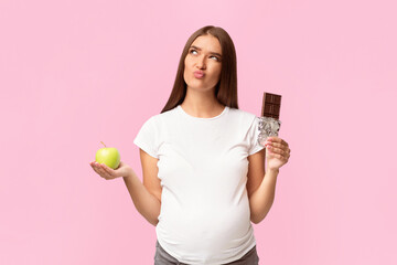 Puzzled Pregnant Woman Choosing Between Chocolate And Green Apple Standing In Studio On White Background. Pregnancy And Nutrition.