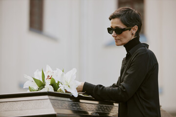 Senior widow in mourning clothes adjusting white lilies on black coffin next to church