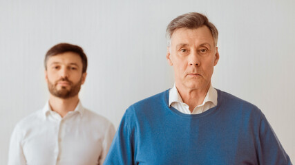 Serious Senior Man And His Middle Aged Son Standing Looking At Camera Against White Wall. Panorama, Selective Focus