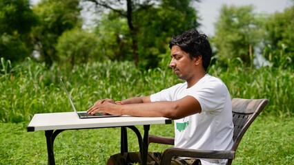 Indian young boy doing work from home. Man sitting on office table at garden doing work on internet