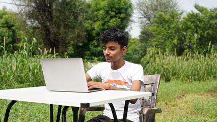 Indian young boy doing work from home. Man sitting on office table at garden doing work on internet