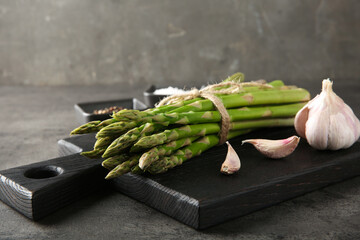 Board with bunch of fresh green asparagus stems and garlic on grey textured table, closeup