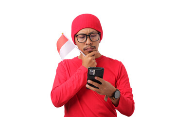 Confused young Indonesian man in a red shirt looks at his phone thoughtfully while holding the Indonesian flag during Indonesia Independence Day celebration on August 17, isolated on white background