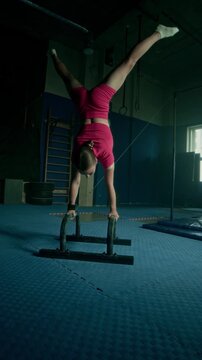 Vertical lowkey shot of hard working Caucasian girl doing perfect handstand on two parallel bars while training alone at dark gym