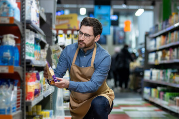 Store employee kneeling in supermarket aisle arranging products. Man wearing apron and glasses focused on organizing items on shelf. Busy retail setting with shelves stocked with various goods.