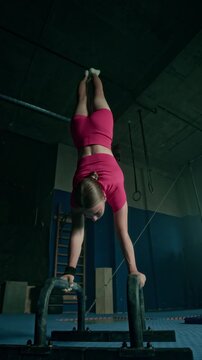 Vertical lowkey shot of determined young Caucasian woman in pink sportswear practicing handstand exercise on parallel bars at gym