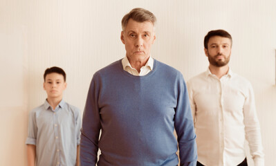 Serious Grandfather With Son And Grandson Looking At Camera Standing Against White Wall Indoor. Selective Focus