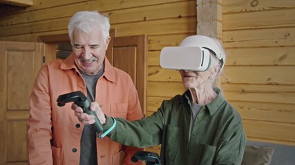 Medium shot of senior woman learning to play augmented reality game with VR goggles and controllers, while chatting to husband, who is teaching and giving advice - Powered by Adobe