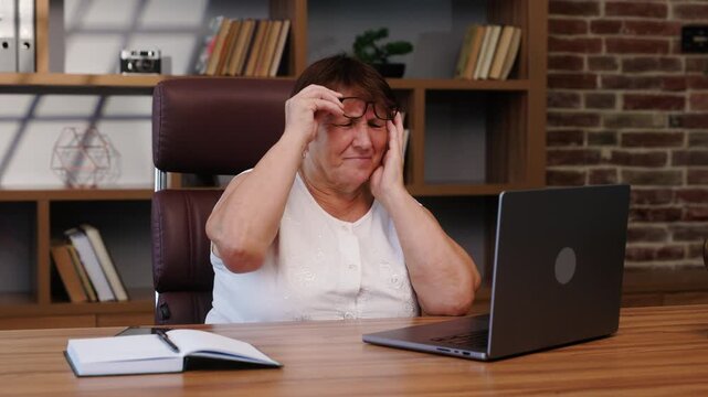 Elderly woman with glasses sitting at a desk in an office, working intently on a laptop. Suddenly, female feels a headache, takes off her glasses, and winces from the migraine.