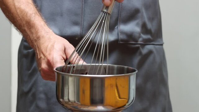 Man in grey apron with whisk and saucepan at kitchen, closeup photo of male hands. Man making chocolate. 