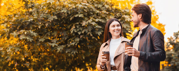 Happy young man and woman having conversation and drinking coffee while spending time at park, empty space