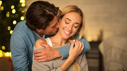 Man kissing neck of stunning woman, sitting near Christmas tree and enjoying time together