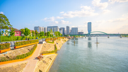 Visitors enjoy a sunny day along the Sava Promenade, with modern buildings in the background and a...