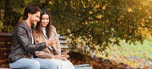 Cheerful guy showing his girlfriend new mobile app while having date at public park, sitting on bench, drinking coffee and looking at smartphone screen, free space