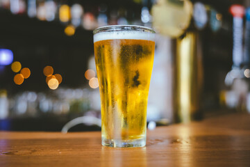 A close-up of a frothy, cold pint of golden beer in a transparent glass, set on a wooden table in a dimly lit pub, evoking a vintage, celebratory atmosphere.