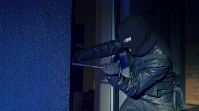 A masked thief breaks down the door of a house at night.