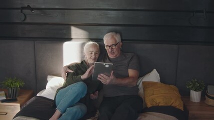 Medium full shot of happy senior Caucasian couple lying on bed together at weekend, hugging, using tablet computer while browsing internet or shopping online, smiling, talking - Powered by Adobe
