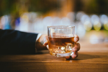 A stylish man holds a glass of whiskey in a fancy pub amidst a crowd of celebrants. The warm and elegant atmosphere accentuates the rich amber-colored liquid.