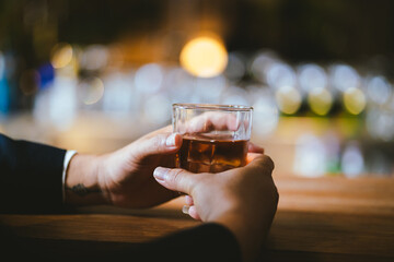A stylish man holds a glass of whiskey in a fancy pub amidst a crowd of celebrants. The warm and elegant atmosphere accentuates the rich amber-colored liquid.