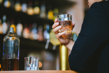 A stylish man holds a glass of whiskey in a fancy pub amidst a crowd of celebrants. The warm and elegant atmosphere accentuates the rich amber-colored liquid.