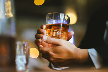 A stylish man holds a glass of whiskey in a fancy pub amidst a crowd of celebrants. The warm and elegant atmosphere accentuates the rich amber-colored liquid.