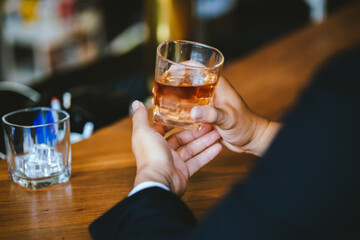 A stylish man holds a glass of whiskey in a fancy pub amidst a crowd of celebrants. The warm and elegant atmosphere accentuates the rich amber-colored liquid.