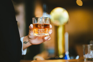 A stylish man holds a glass of whiskey in a fancy pub amidst a crowd of celebrants. The warm and elegant atmosphere accentuates the rich amber-colored liquid.