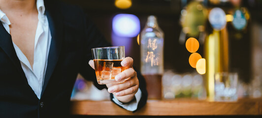 A stylish man holds a glass of whiskey in a fancy pub amidst a crowd of celebrants. The warm and elegant atmosphere accentuates the rich amber-colored liquid.
