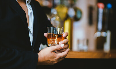 A stylish man holds a glass of whiskey in a fancy pub amidst a crowd of celebrants. The warm and elegant atmosphere accentuates the rich amber-colored liquid.