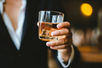 A stylish man holds a glass of whiskey in a fancy pub amidst a crowd of celebrants. The warm and elegant atmosphere accentuates the rich amber-colored liquid.