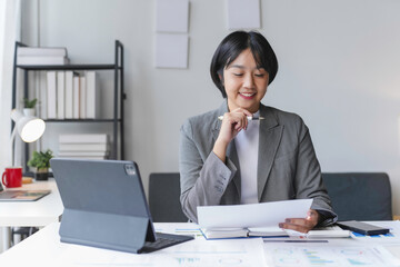 Asian businesswoman in a gray business suit smiles as she reviews documents at her desk