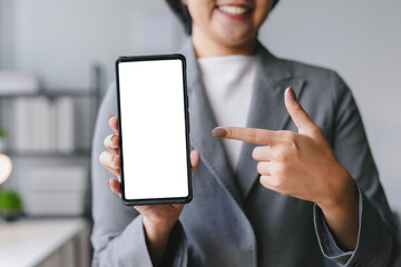 Smiling businesswoman is holding smartphone with white screen in office and pointing at it with index finger