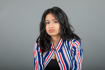 Portrait of sad Indian young woman sulking, frowning and looking upset, distressed frustrated face expression, standing gloomy against grey background.