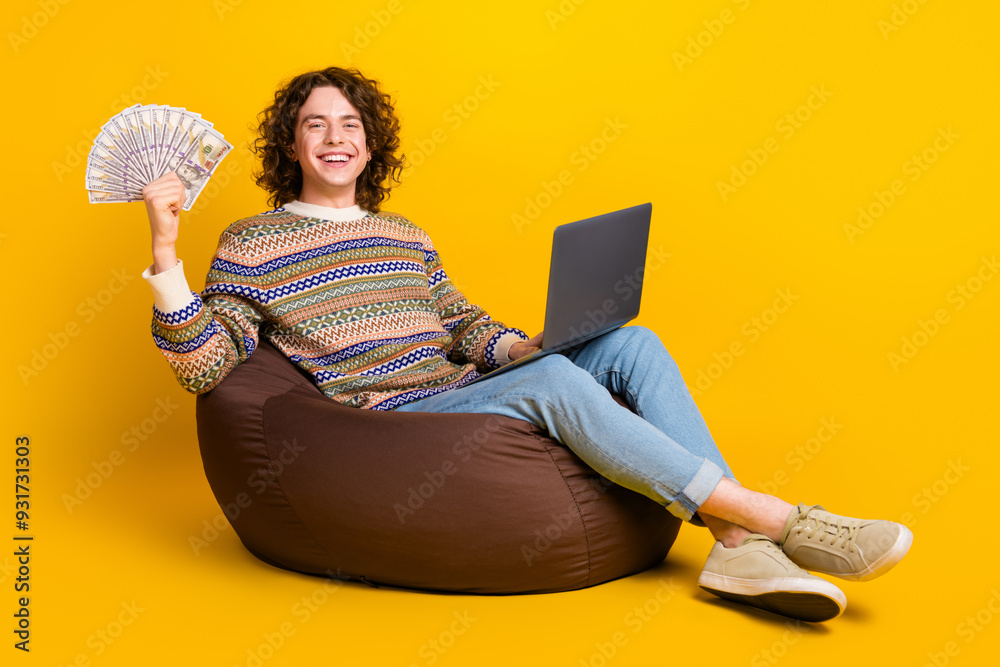 Poster Full size photo of satisfied smart guy dressed sweater sit on pouf with laptop hold bunch of money isolated on yellow color background