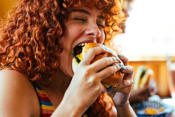 Multiethnic group of friends having meal at 80s vintage diner restaurant, woman biting cheeseburger...